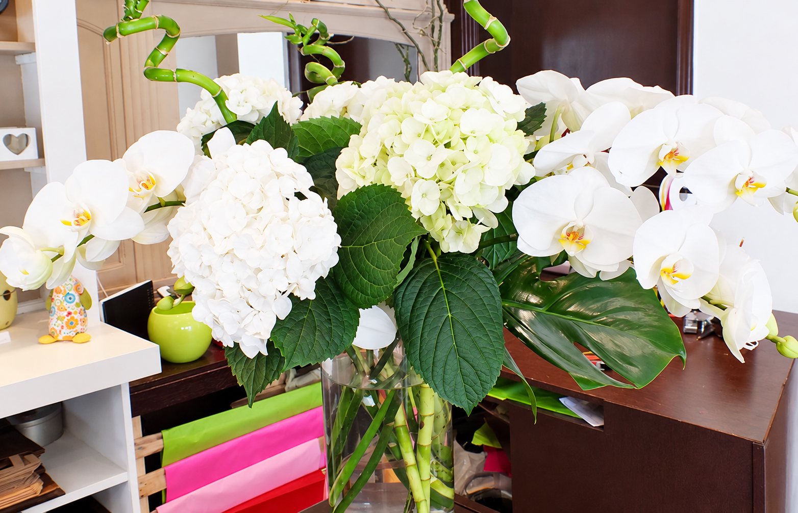 Un grand bouquet d'hortensias blancs et d'orchidées phalaenopsis dans un vase en verre, posé dans une boutique de fleuriste avec du papier de soie coloré en dessous.