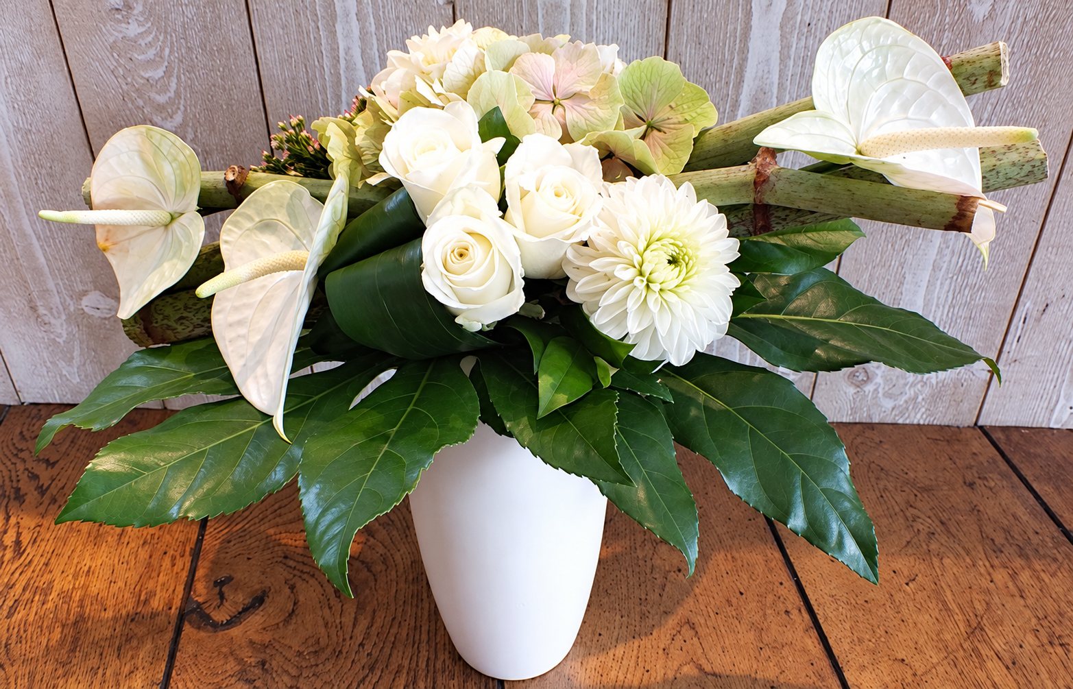 Bouquet composé de roses, dahlia, hortensia et anthuriums blancs entourés de feuillage vert, posé dans un vase blanc sur une table en bois devant un mur en planches grisées.