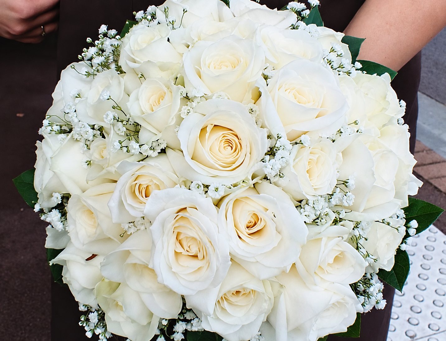 Une personne tient un bouquet rond composé de roses blanches crème, de gypsophile et de feuilles vertes.