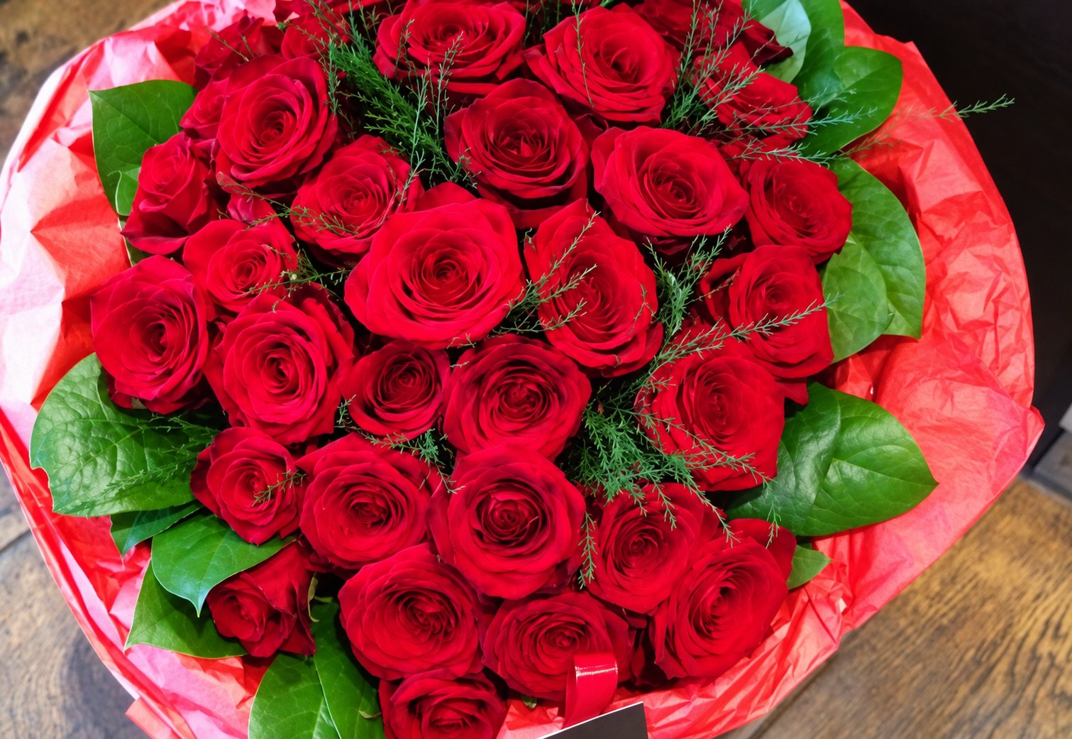 Vue de dessus d'un bouquet rond de roses rouges entouré de feuilles vertes et de papier d'emballage rouge et rose, posé sur une surface en bois.