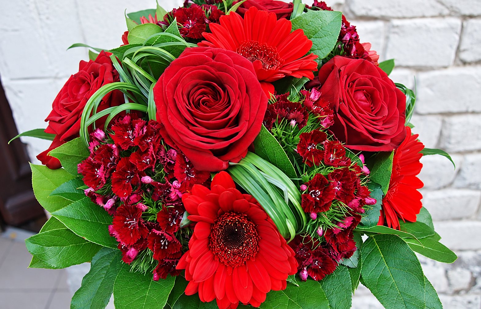 Gros plan d'un bouquet rond composé de roses rouges, de gerberas rouges, de petits œillets sombres et de feuilles vertes devant un mur de briques blanchi.
