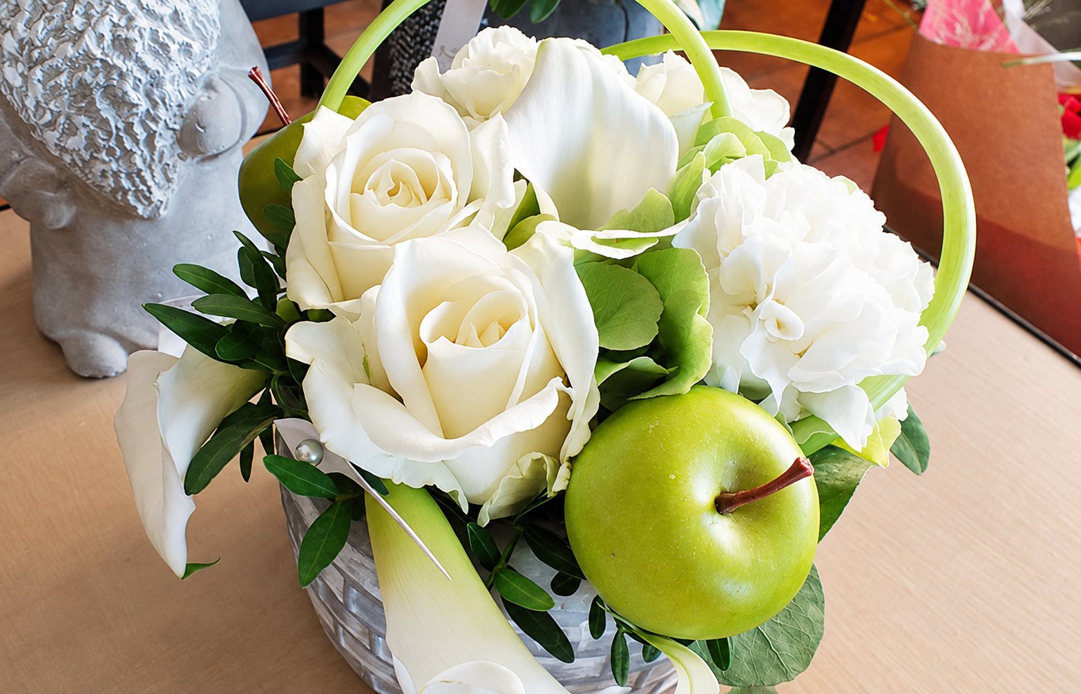 Composition florale de roses blanches, d'œillets et de callas avec une pomme verte, posée sur une table près d'une statuette en pierre.