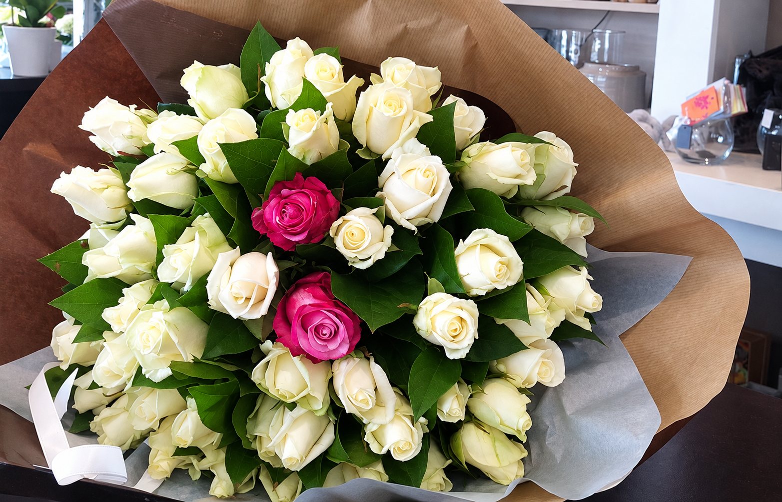 Un bouquet de roses blanches avec deux roses roses au centre, emballé dans du papier kraft et du papier gris, posé dans une boutique.