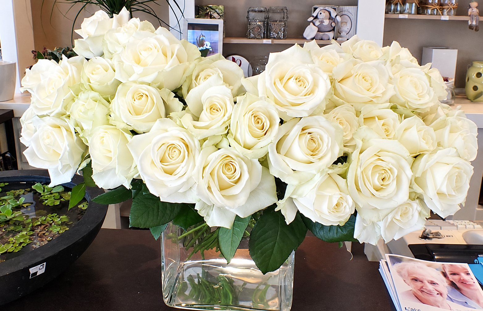 Un grand bouquet de roses blanches dans un vase en verre carré posé sur un comptoir sombre, avec des étagères décorées en arrière-plan.