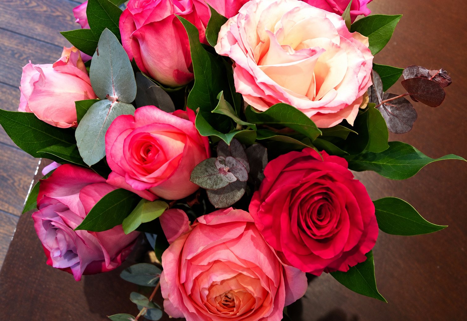 Vue de dessus d'un bouquet de roses roses, rouges et pêche mêlées à des feuilles vertes et à de l'eucalyptus, posé sur une table en bois.