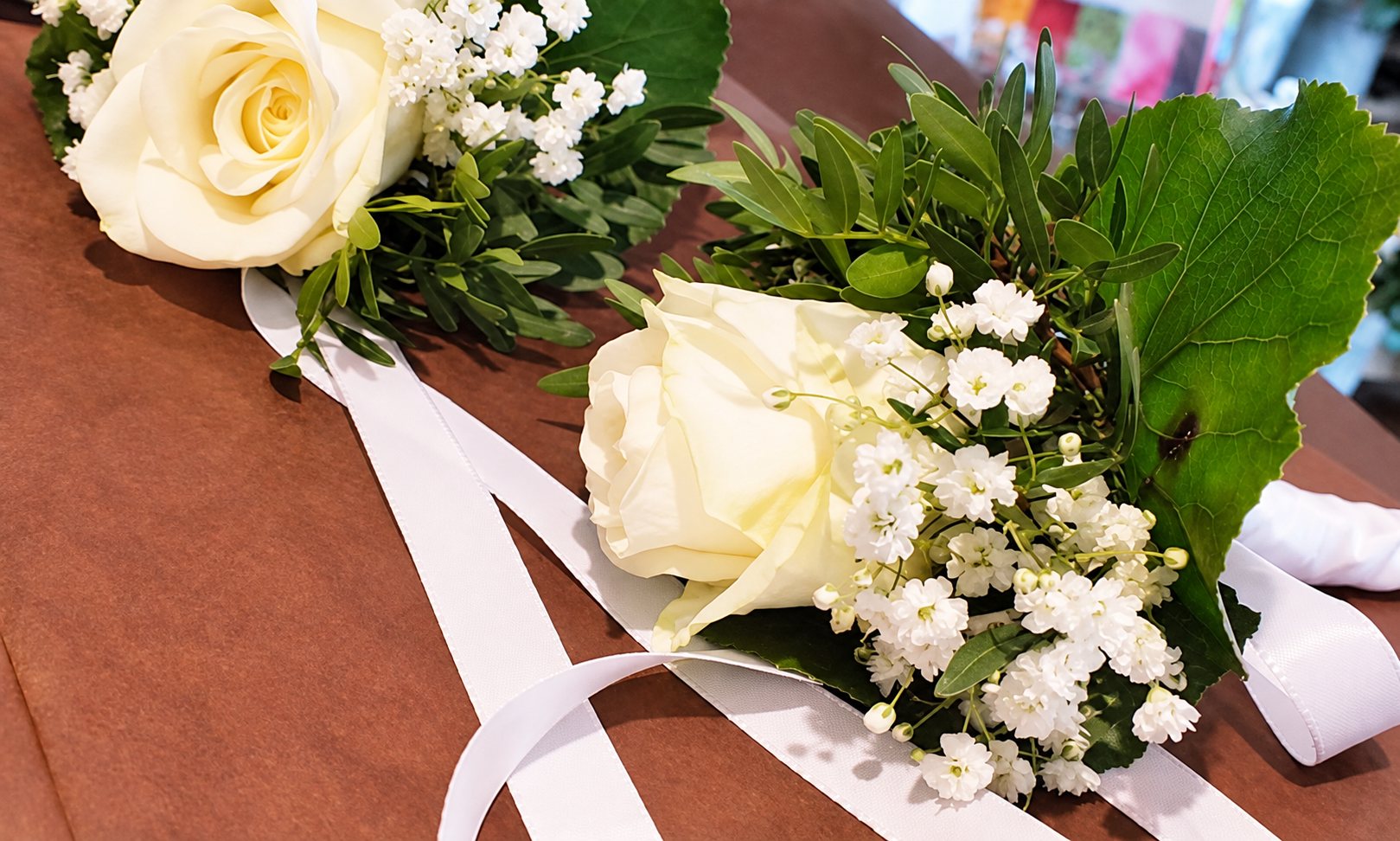 Deux boutonnières composées de roses blanches, de gypsophile et de feuillage, posées avec des rubans blancs sur du papier kraft brun.