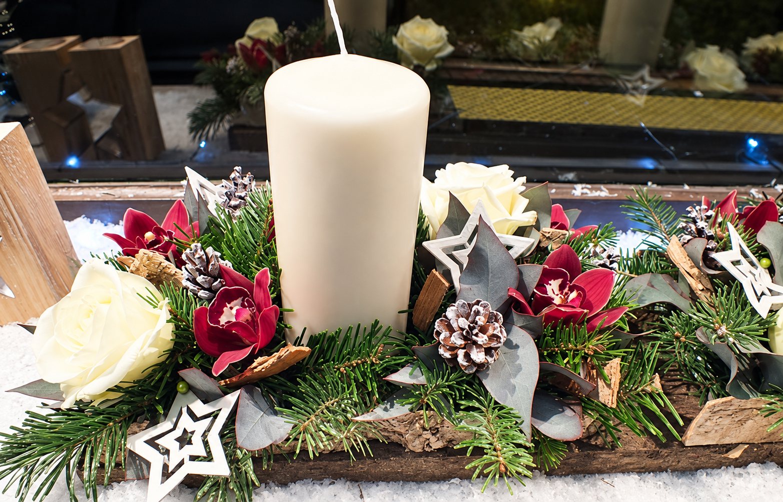 Composition florale de Noël sur un support en bois, avec une grande bougie blanche, des orchidées bordeaux, des roses blanches, des branches de sapin, des pommes de pin et des étoiles décoratives.