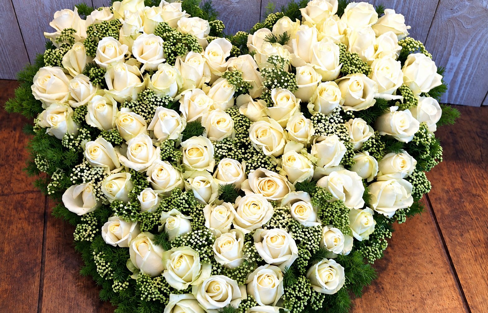Un grand cœur de roses blanches entouré de verdure et de petites fleurs, posé sur une table en bois.