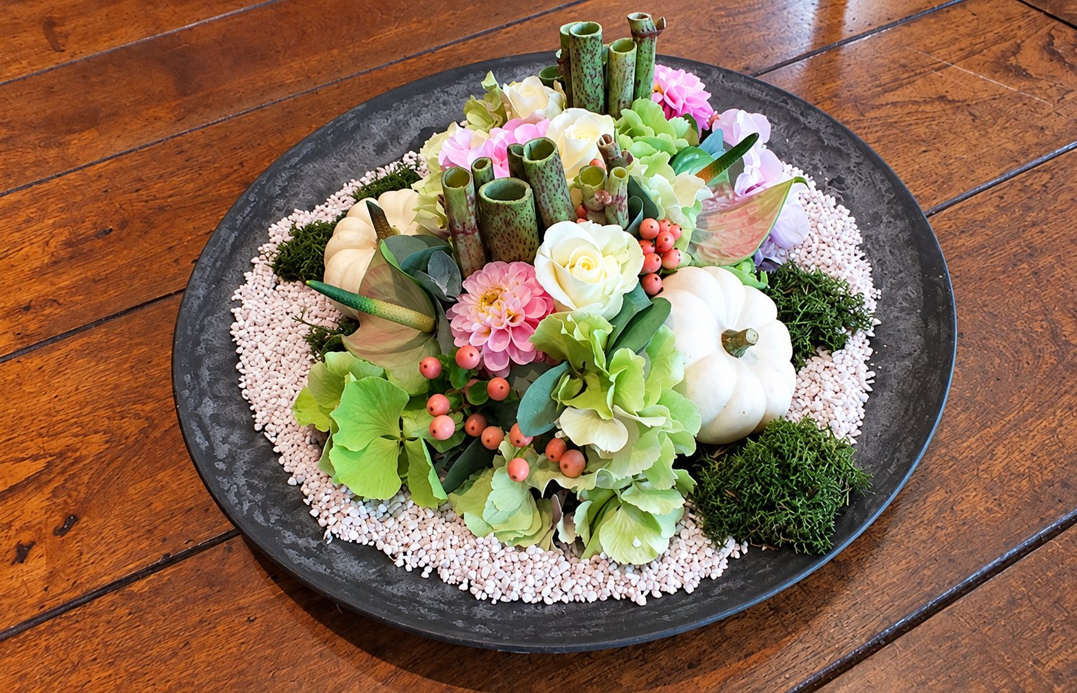 Composition florale dans un grand plat gris avec roses, dahlias roses, hortensias verts, petites citrouilles blanches, mousse et gravier clair, posée sur une table en bois.