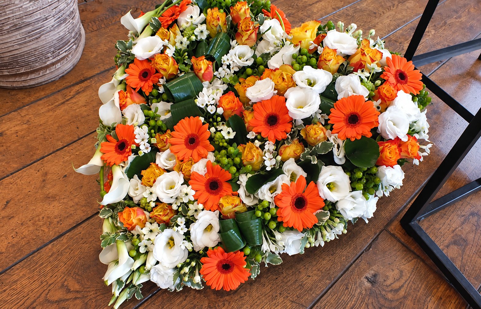 Une composition de fleurs en forme de cœur, mêlant gerberas orange, roses orange et lisianthus blancs, posée sur un parquet en bois.