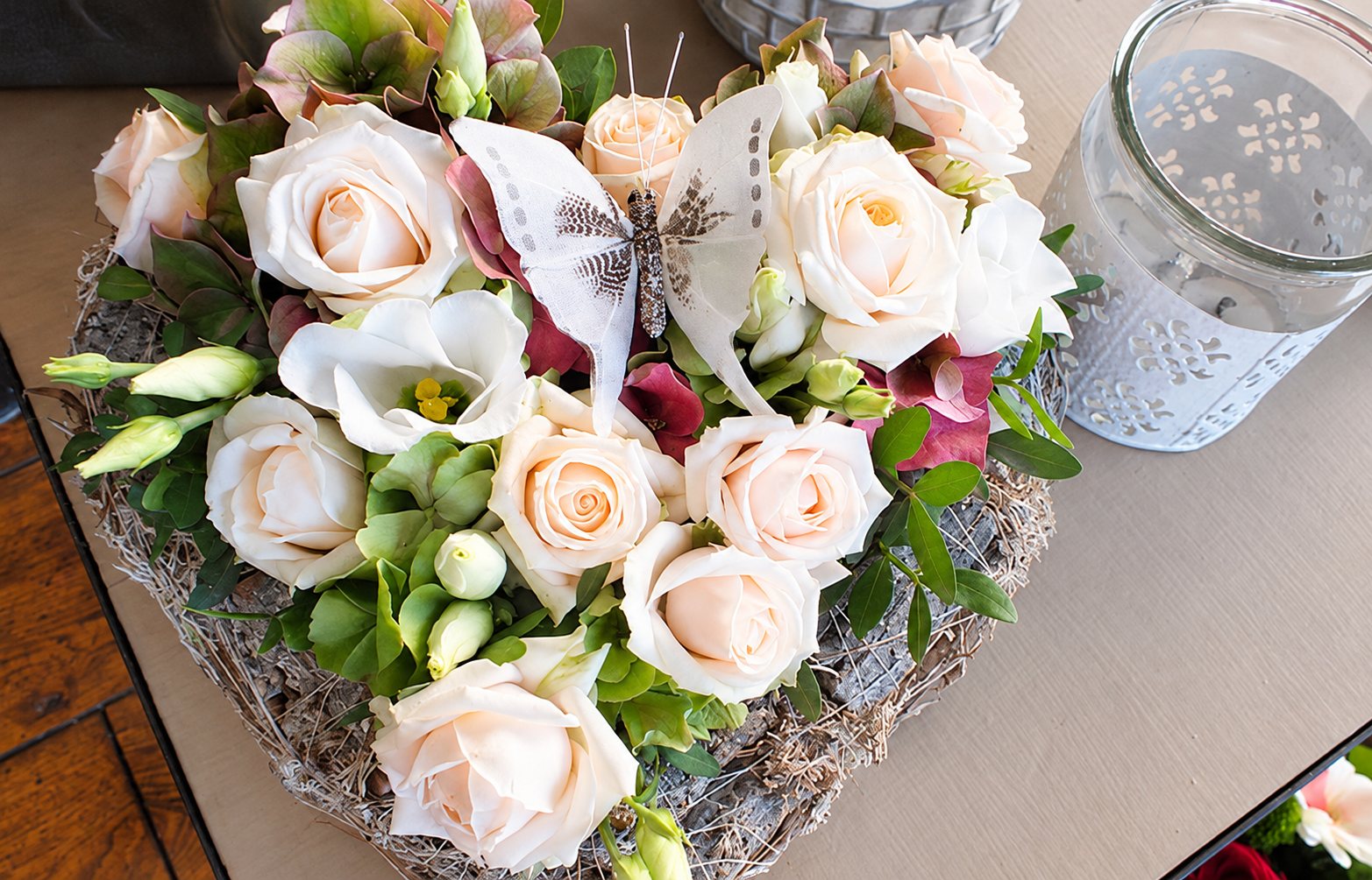 Une composition de roses couleur pêche, de lisianthus et de feuillage dans un support en forme de cœur, ornée d'un papillon décoratif, posée sur une table à côté d'un photophore en verre.