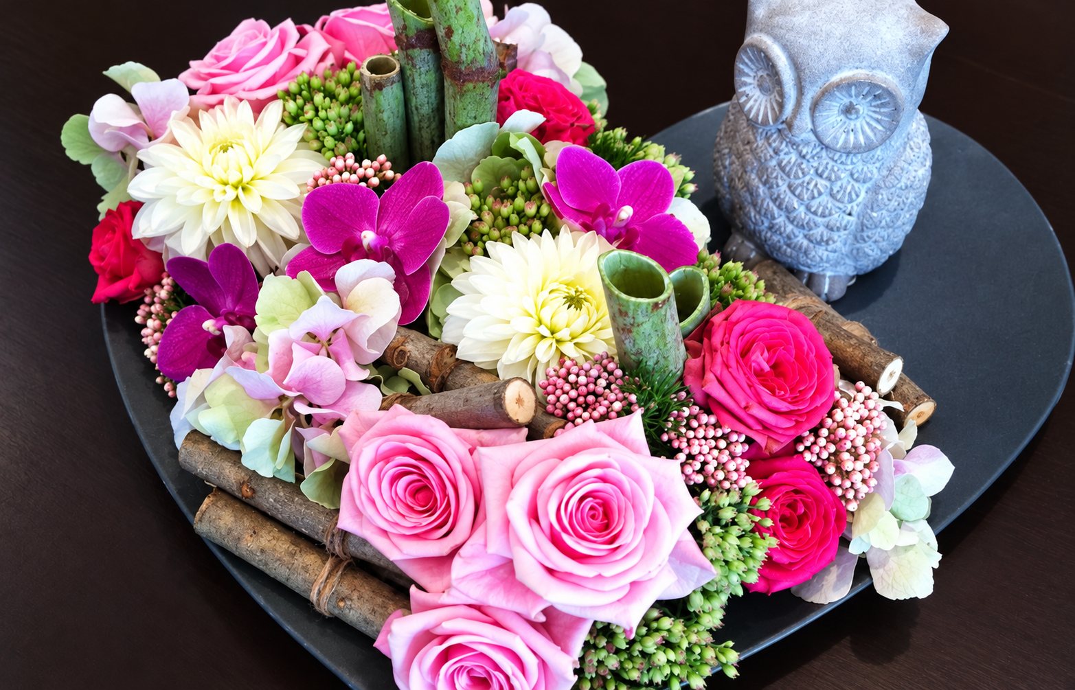 Composition de roses roses, d'orchidées et de dahlias blancs disposée sur un plateau en forme de cœur, avec une petite chouette bleu pâle.