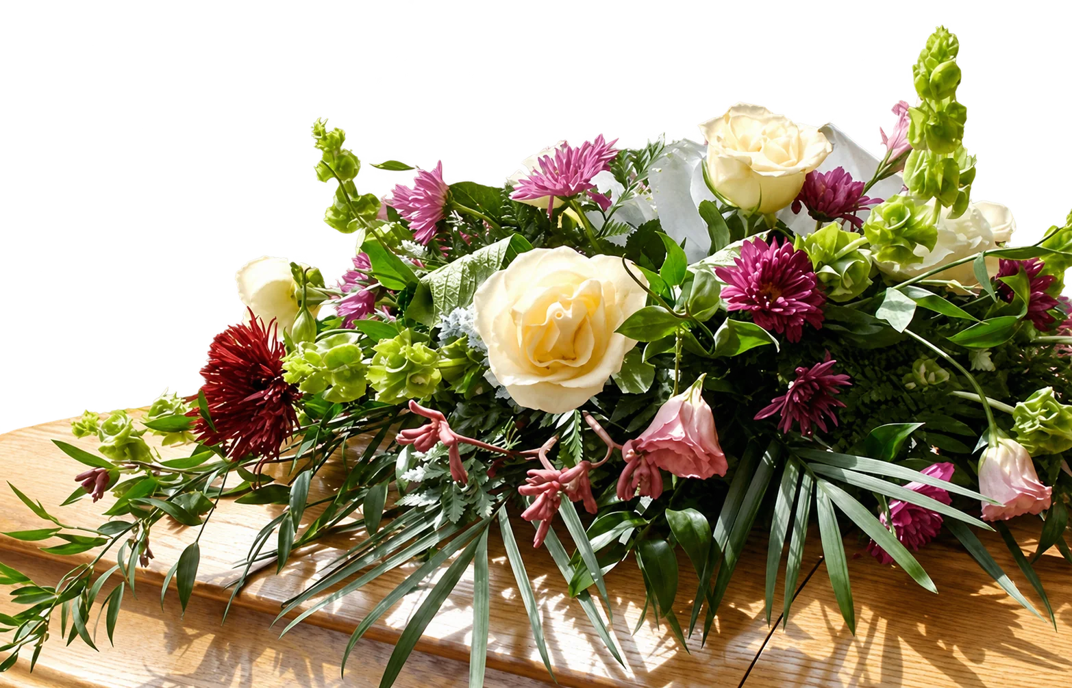 Une gerbe de fleurs composée de roses blanches, de chrysanthèmes violets et de feuillages verts est posée sur un cercueil en bois clair.