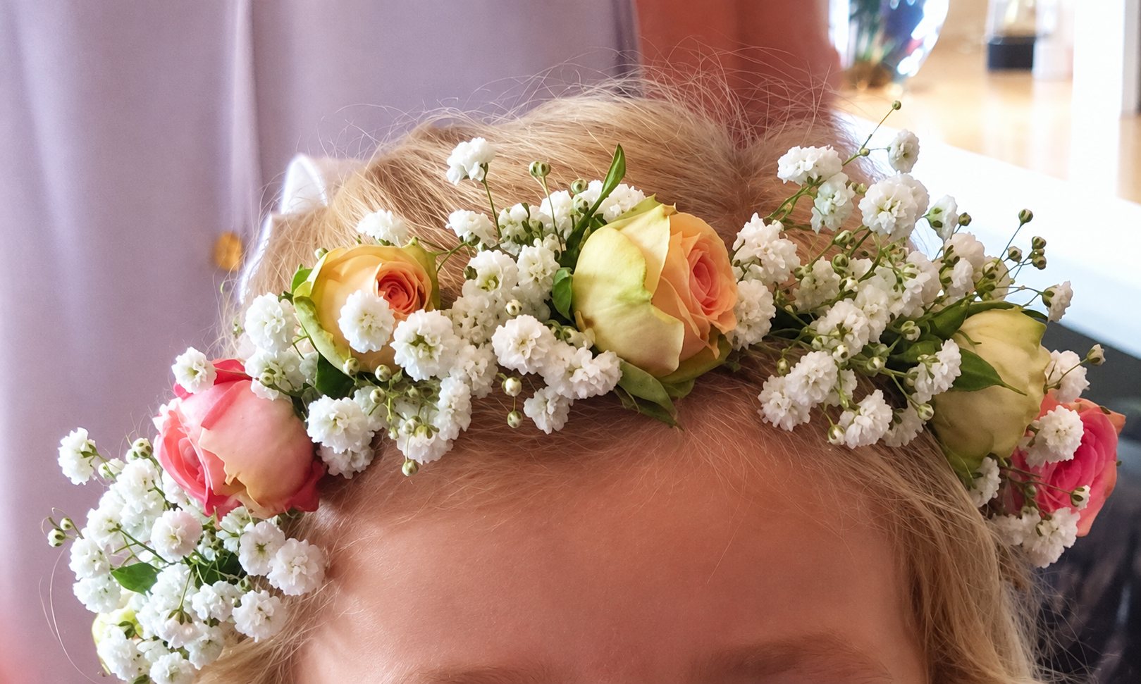 Gros plan sur une couronne de fleurs composée de gypsophile blanche et de petites roses roses, jaunes et orangées posée sur les cheveux blonds d'une personne.