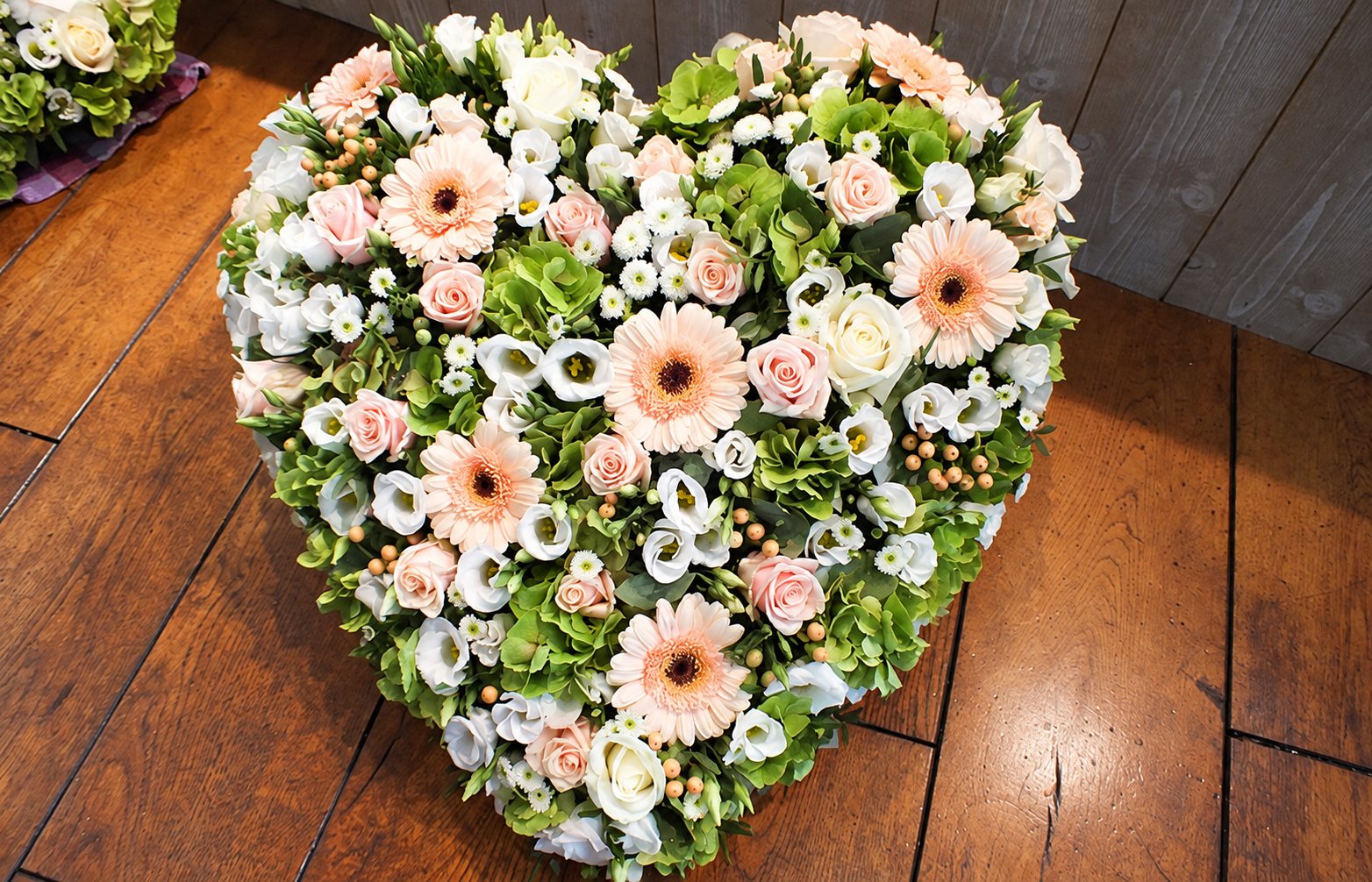 Composition florale en forme de cœur mêlant gerberas pêche, roses, lisianthus blancs et hortensias verts, posée sur un parquet en bois.