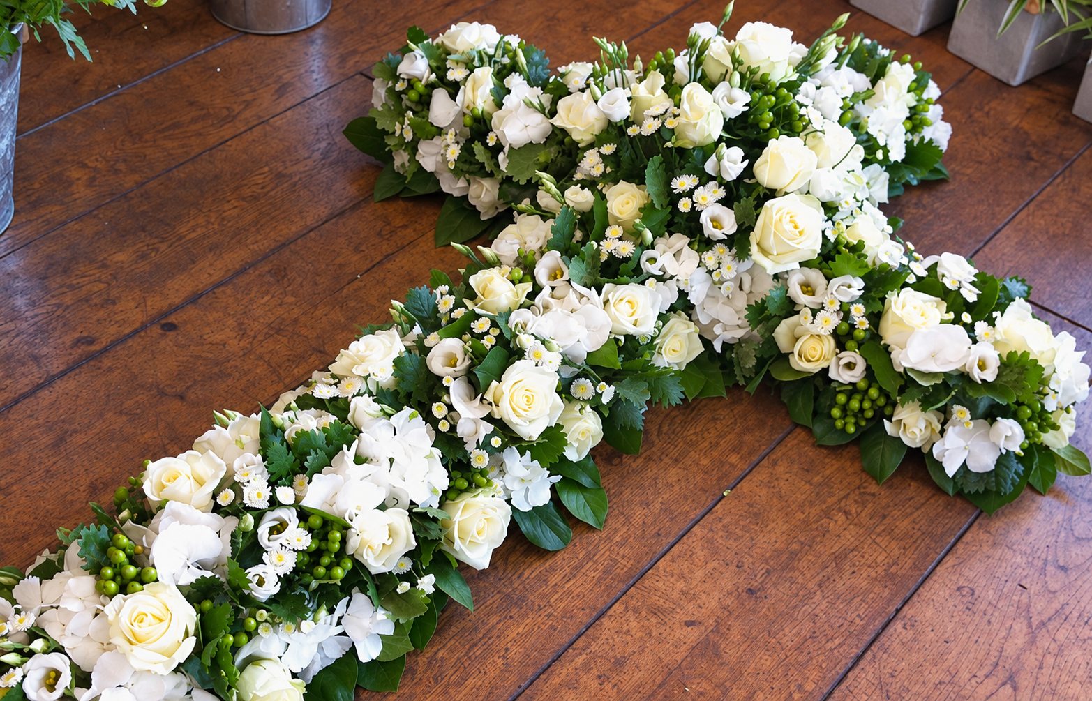 Une grande croix de fleurs composée de roses blanches, de petites fleurs blanches et de feuillage vert, posée sur un parquet en bois foncé.