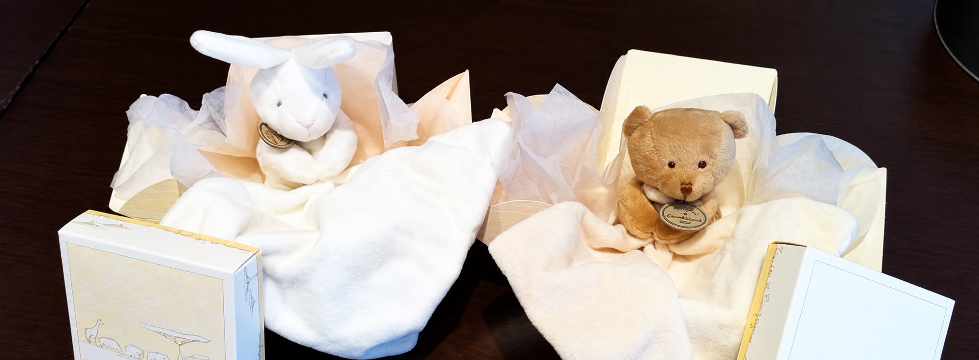 Un doudou lapin blanc et un doudou ourson beige sont présentés sur leurs mouchoirs en tissu, entourés de papier de soie et de boîtes cadeaux crème, posés sur une table sombre.