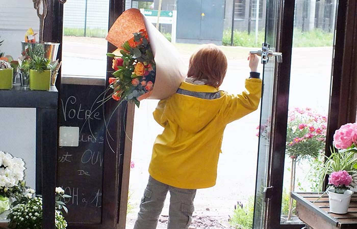 Un enfant en ciré jaune ouvre la porte d'une boutique de fleuriste en tenant un grand bouquet de roses orange emballé de papier kraft.