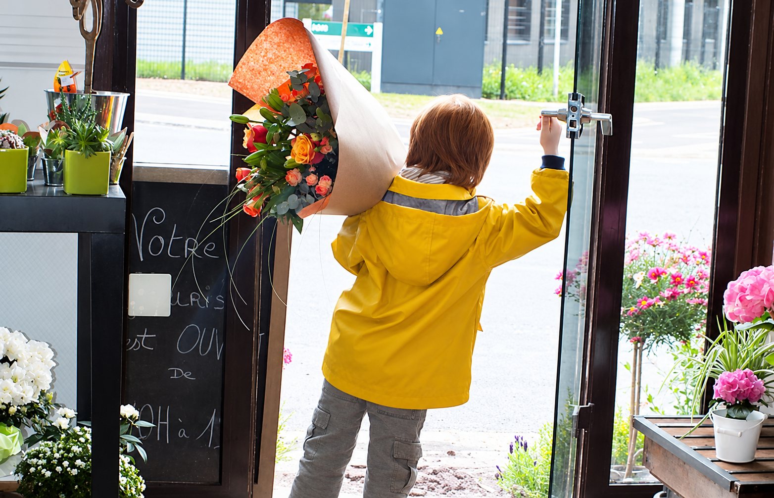 Un enfant en ciré jaune pousse la porte de la boutique en portant un grand bouquet emballé dans du papier kraft
