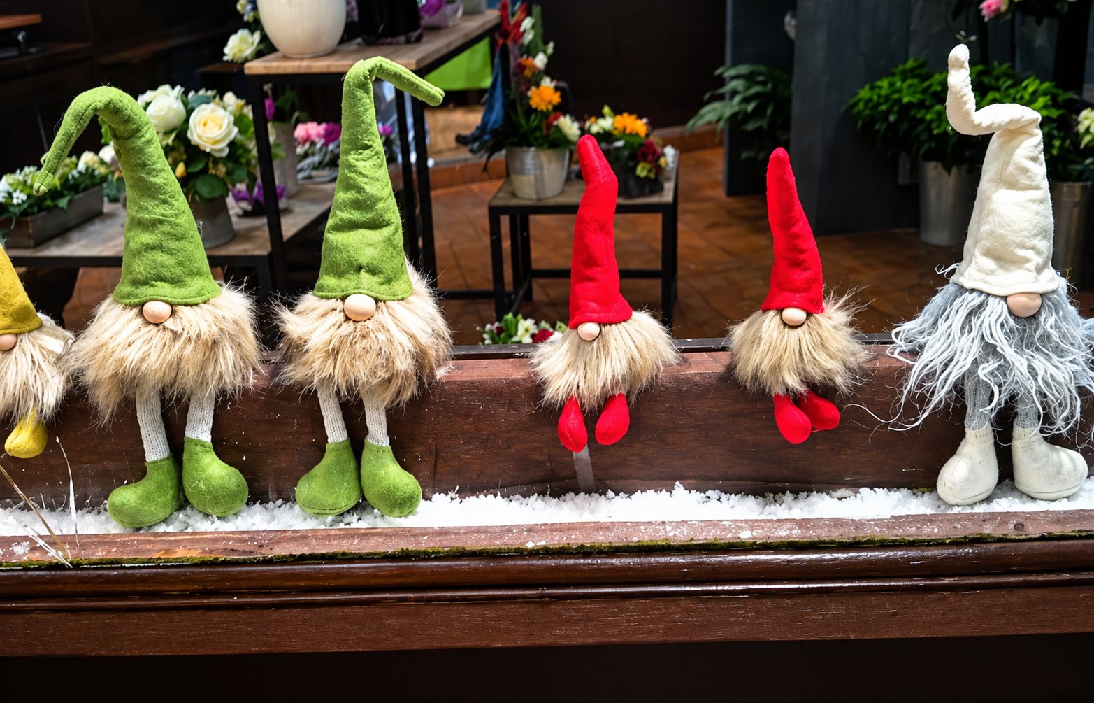 Une rangée de gnomes de Noël en peluche, aux bonnets verts, rouges et crème, assis sur un rebord de vitrine garni de fausse neige devant des bouquets de fleurs.