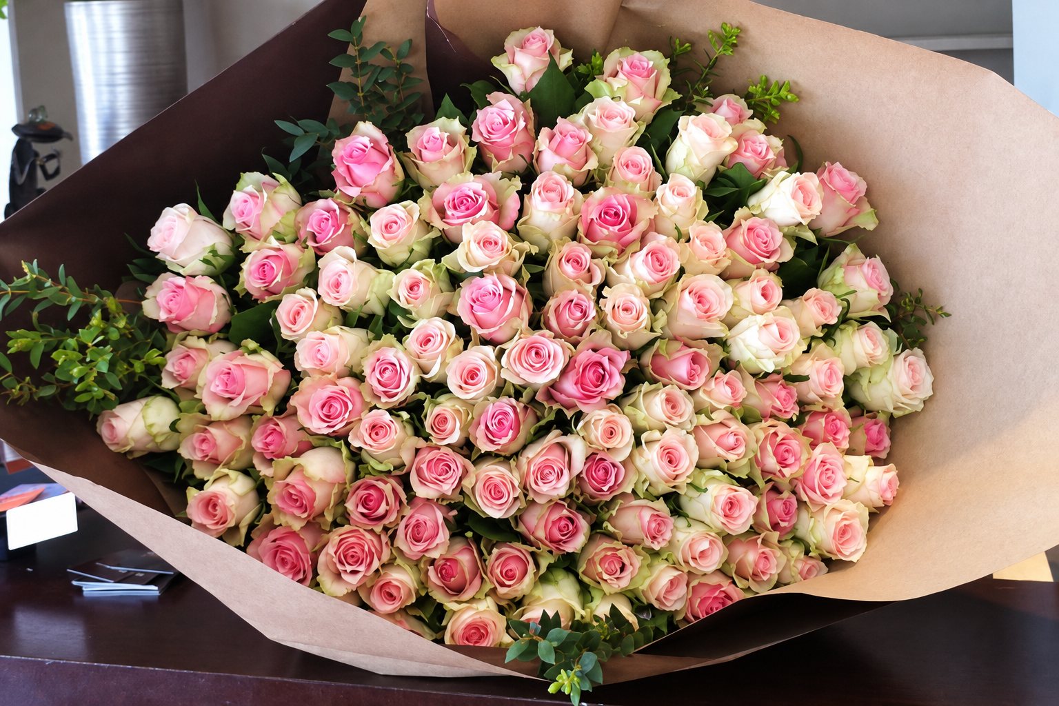 Un très grand bouquet de roses rose pâle et blanches, enveloppé dans du papier kraft et posé sur une table en bois sombre.