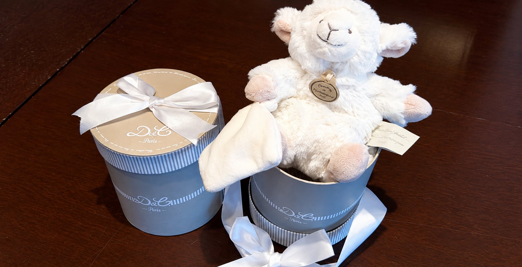 Une peluche d'agneau blanc avec son doudou sort d'une boîte ronde grise à ruban blanc, à côté d'une seconde boîte fermée, posées sur une table en bois sombre.