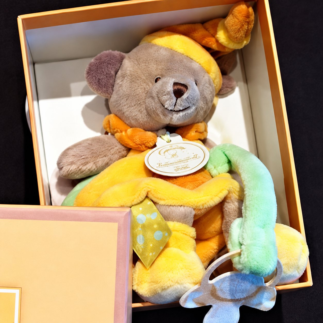 Un doudou ours gris, jaune et vert avec une étiquette et une tétine est installé dans une boîte cadeau beige ouverte.
