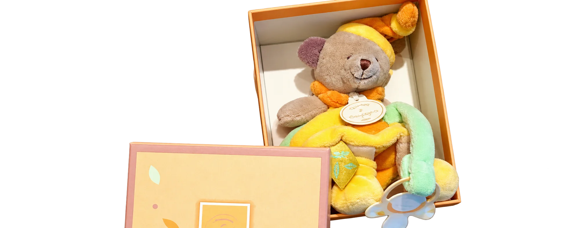 Un ourson en peluche beige et jaune avec un anneau vert est présenté dans une boîte cadeau orange, posée sur un fond sombre à côté de son couvercle.