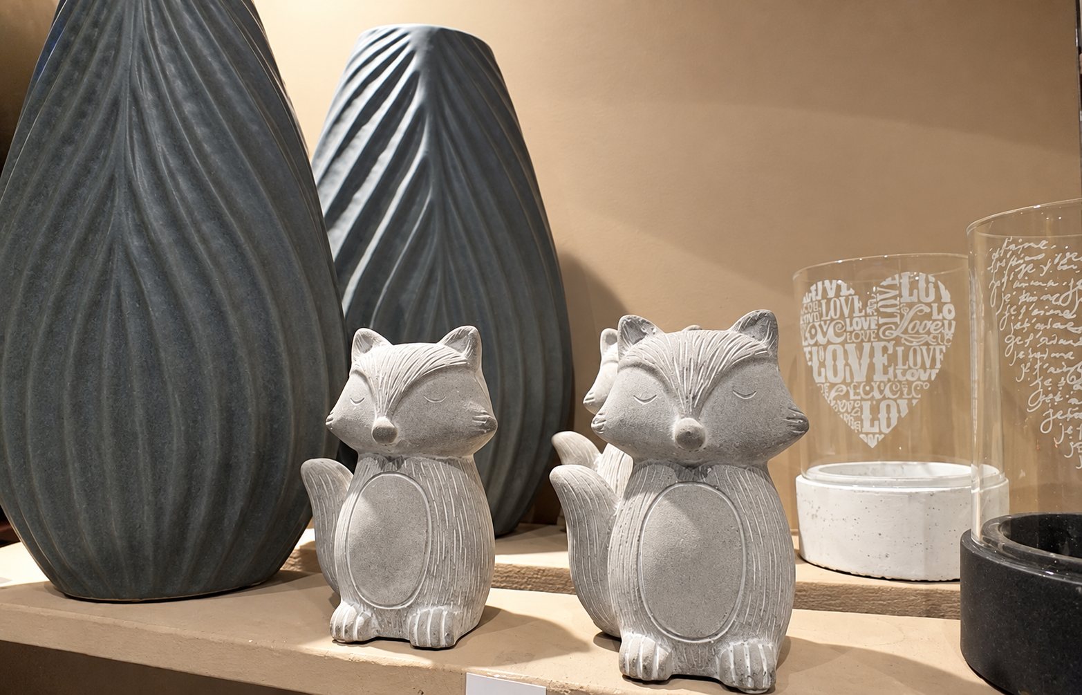 Deux petits renards décoratifs en béton gris posés sur une étagère devant de grands vases nervurés et des photophores en verre ornés de cœurs.